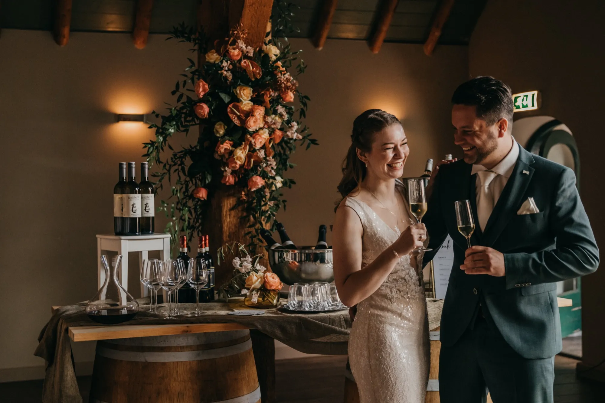 Bruid en bruidegom lachen met een glas champagne bij een bloemenzuil